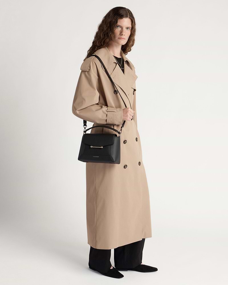 Model in a beige trench coat holding a black mosaic bag with shoulder strap.