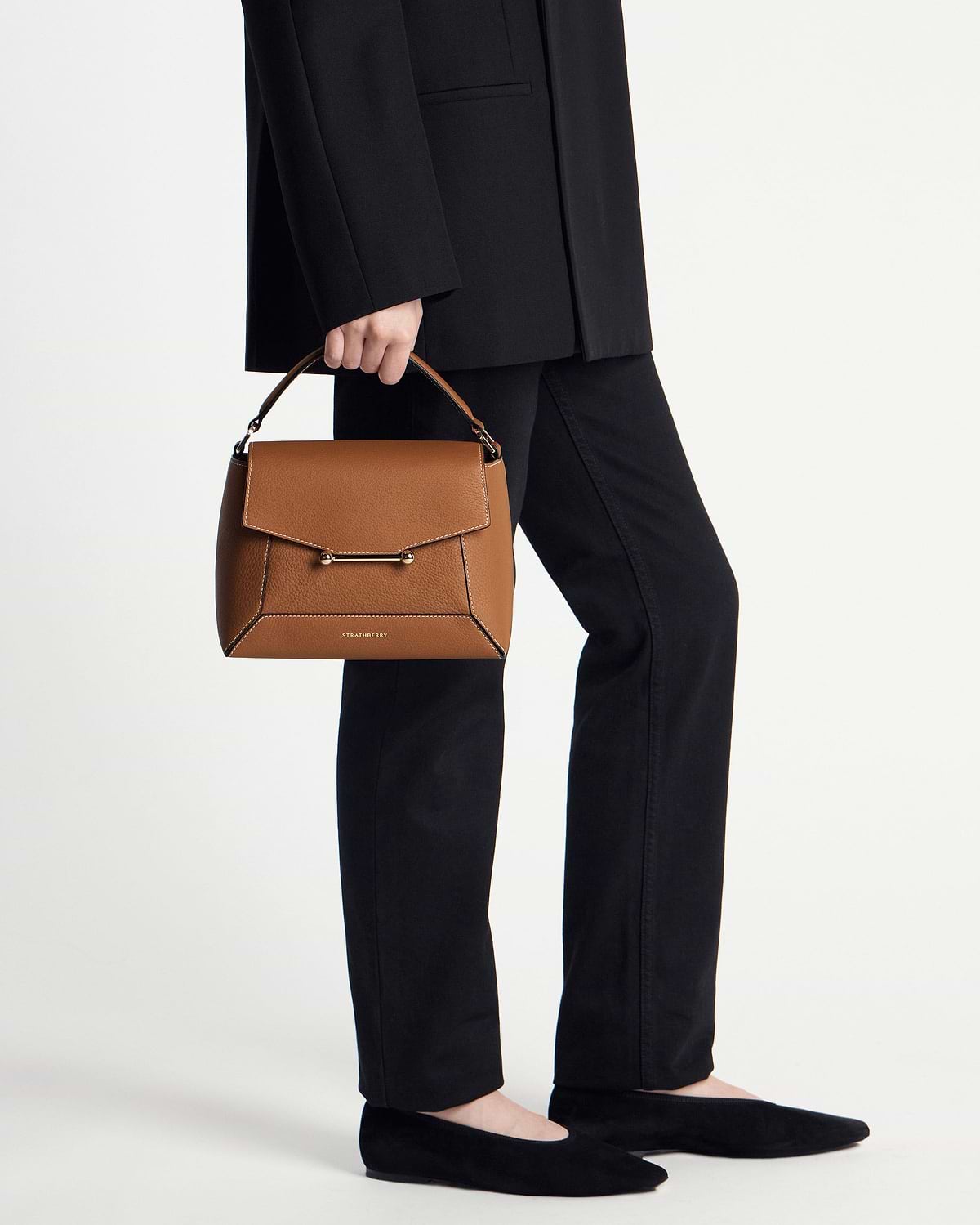 Model cropped below waist holding tan Mosaic bag with vanilla stitching, Strathberry logo visible.