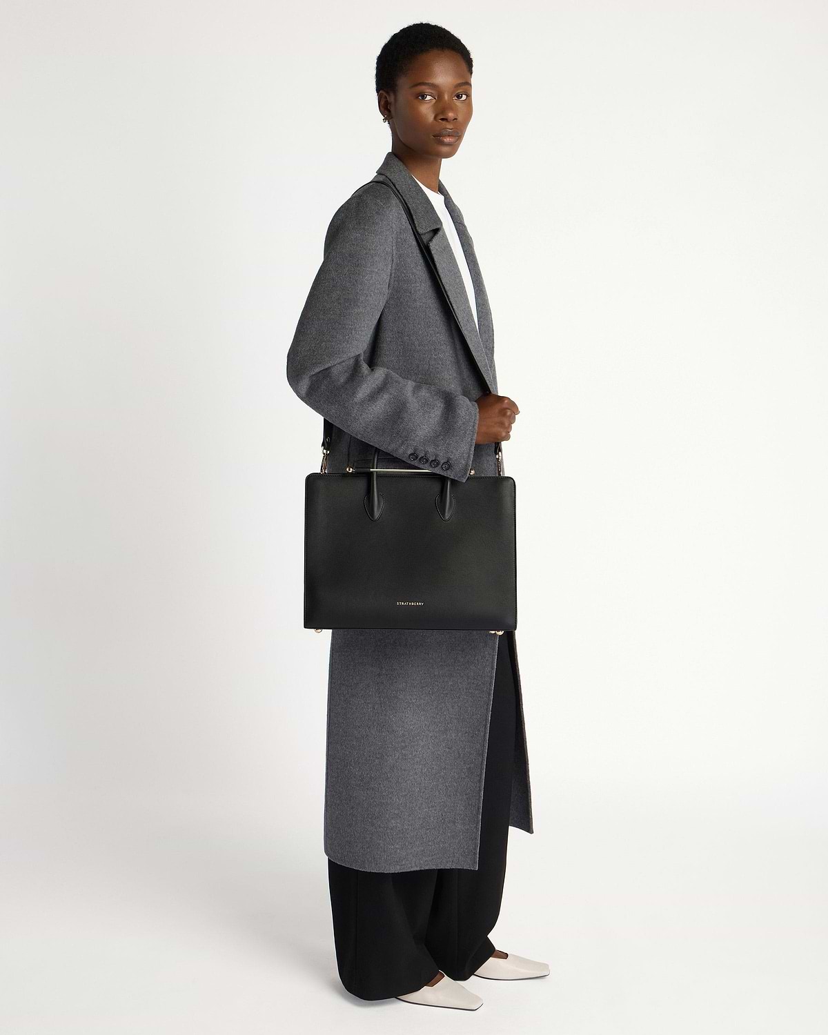 Model wearing grey coat holding black Strathberry tote bag over shoulder.
