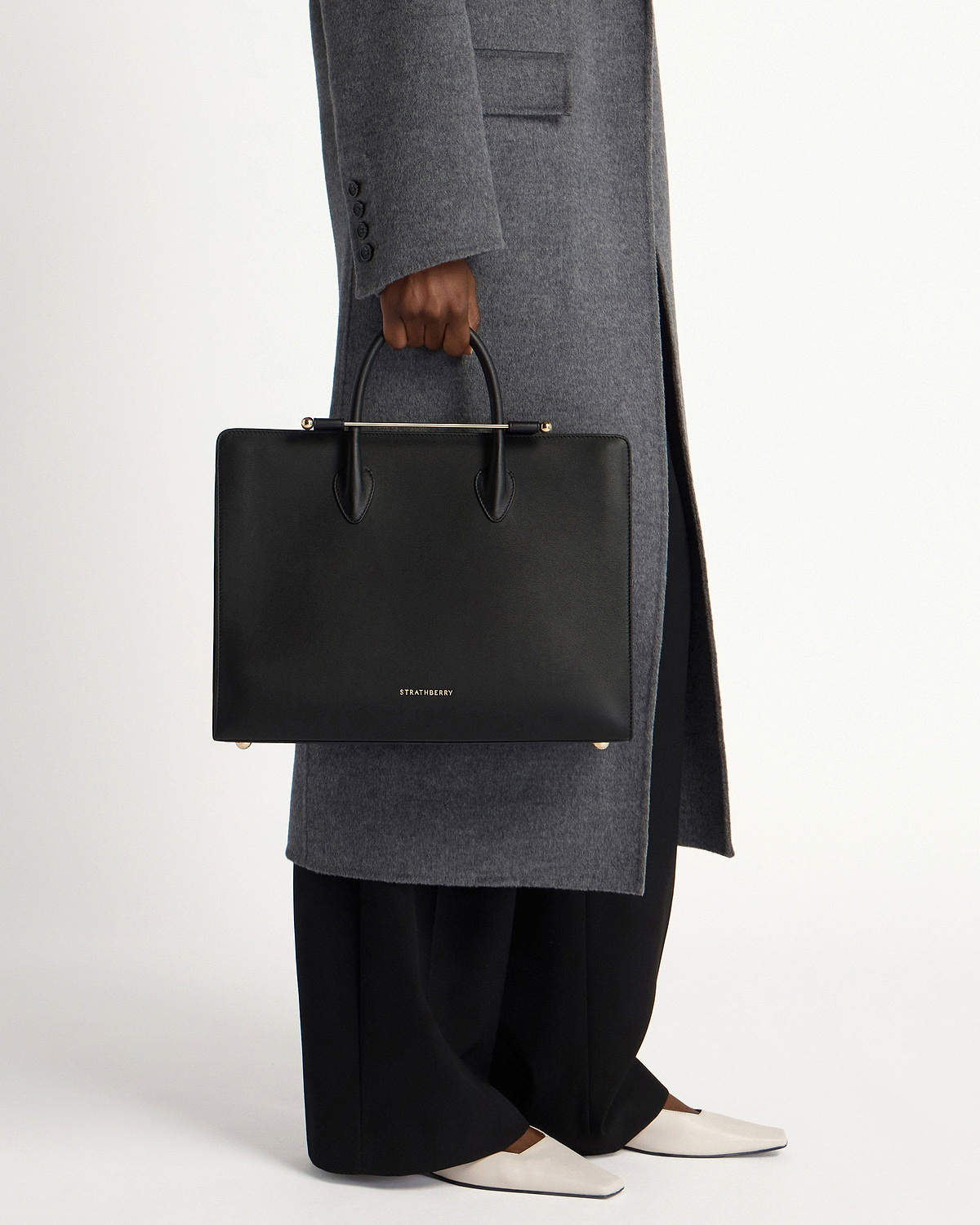 Black Strathberry leather tote with top handles held by model wearing grey coat