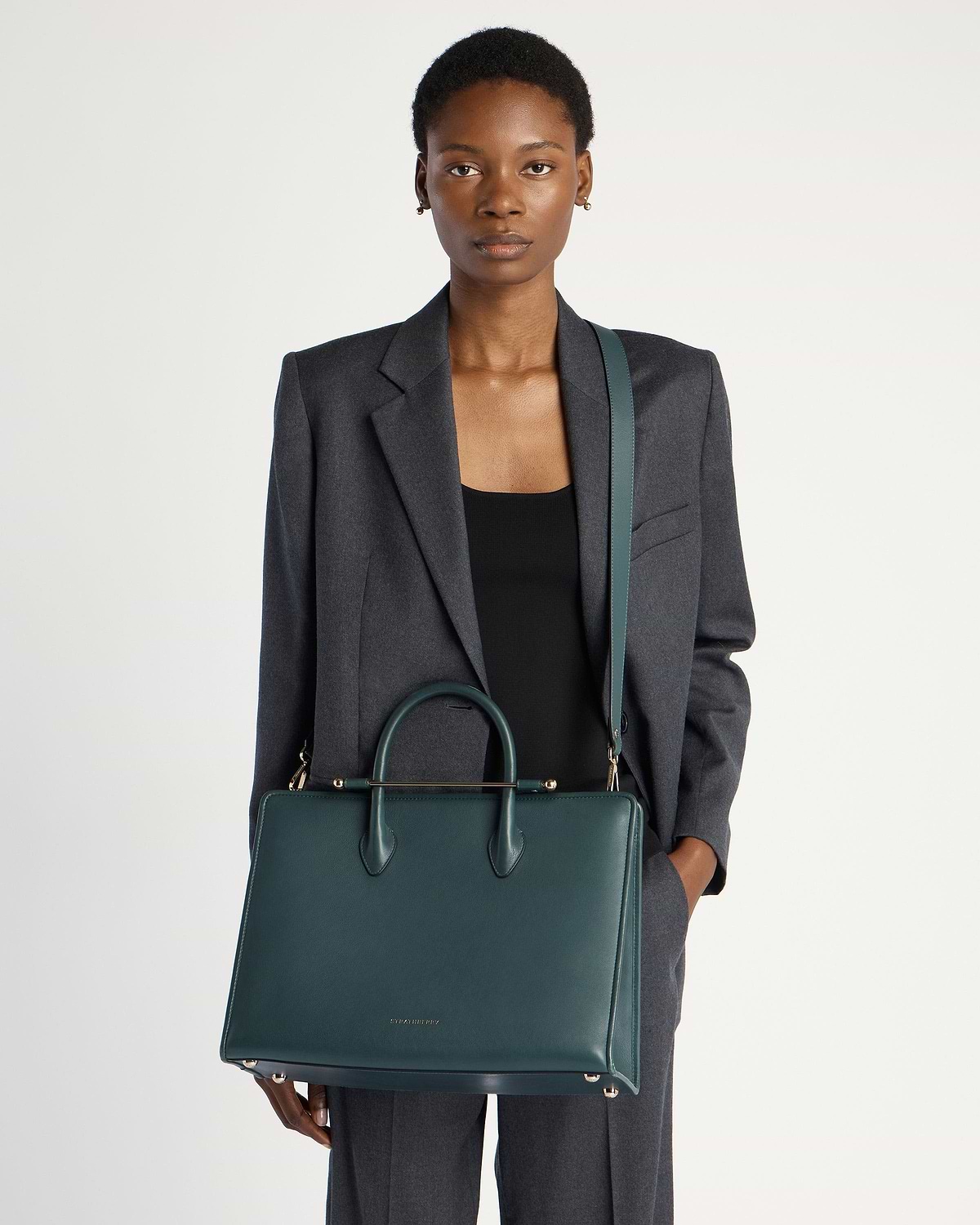 Model wearing a grey blazer holds a bottle-green Strathberry leather tote bag.