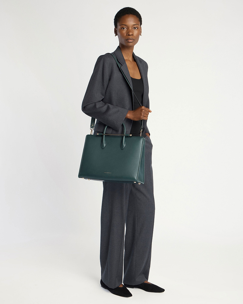 Woman in grey suit standing, holding Strathberry bottle green tote bag with strap.