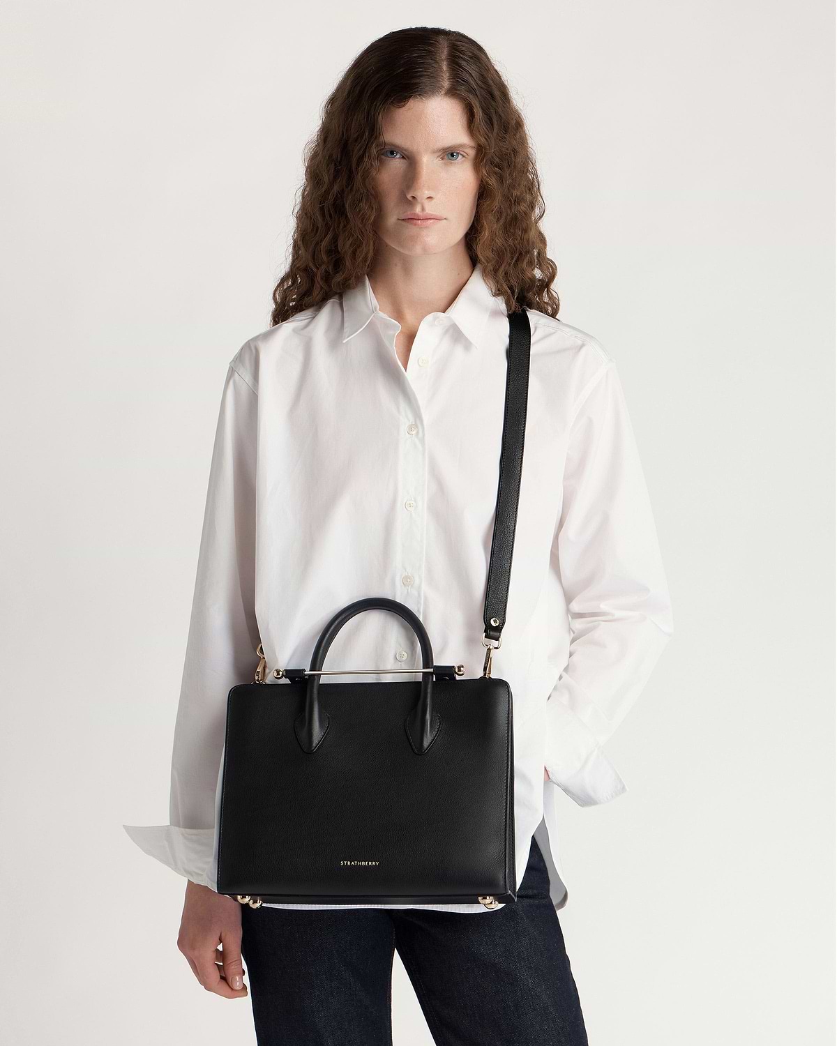 Woman wearing white shirt carrying black Strathberry midi tote with top handles and shoulder strap.