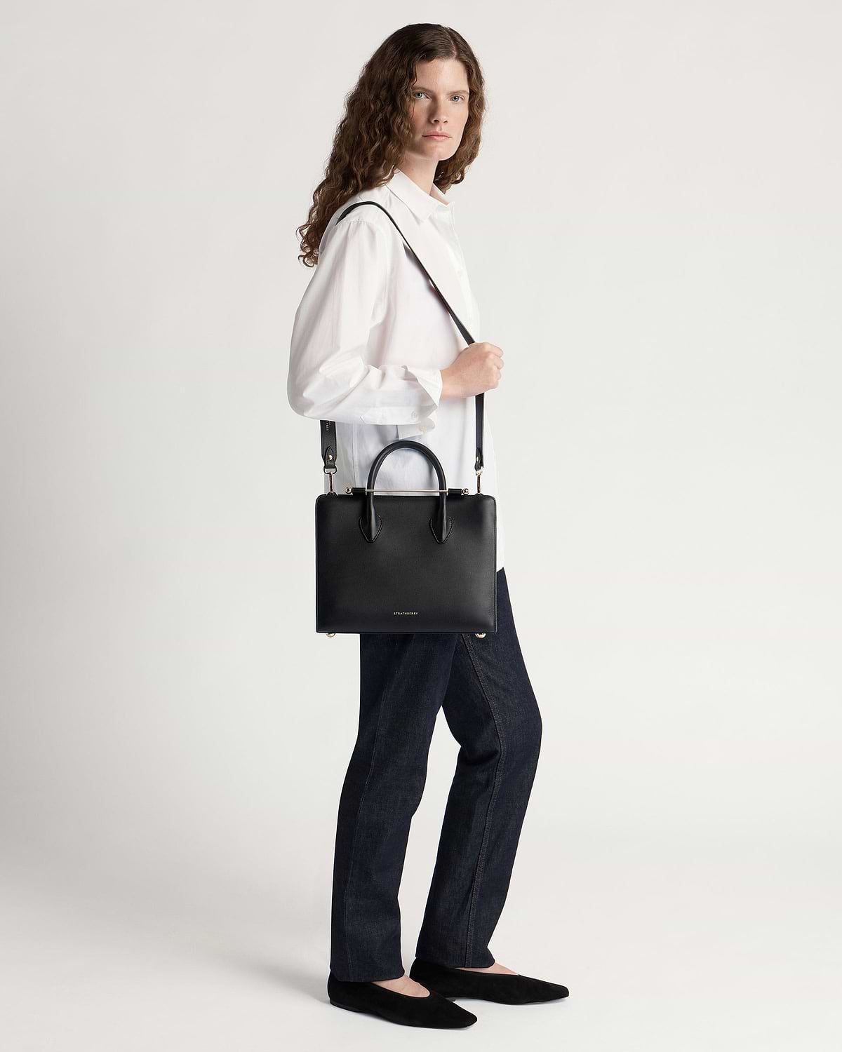 Model wearing a white shirt and jeans carrying a black Strathberry midi tote.