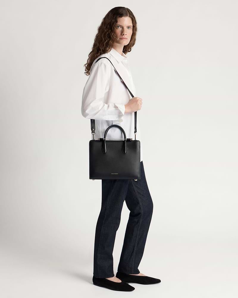 Model wearing a white shirt and jeans carrying a black Strathberry midi tote.