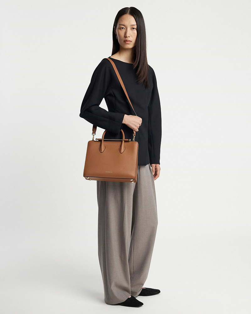 Model wearing black top and grey wide trousers holding chestnut midi tote bag.