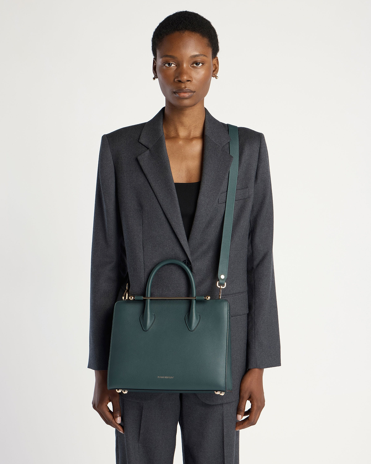 Model wearing grey suit carries bottle green Strathberry midi tote with shoulder strap