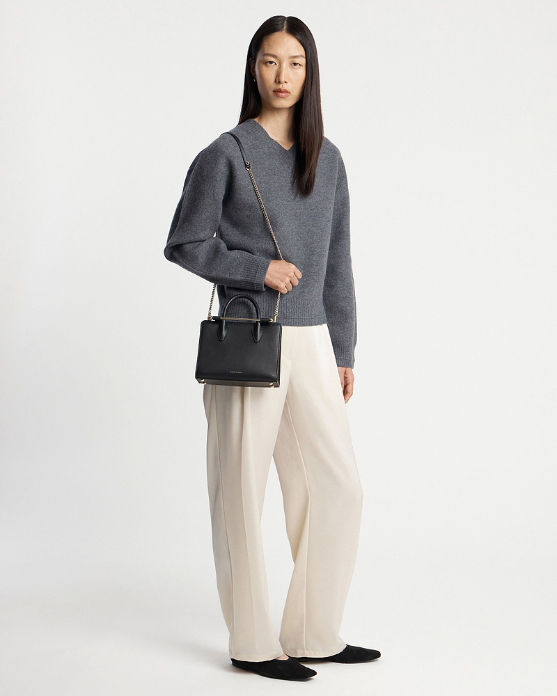 Model wearing grey jumper and cream trousers holding a black mini tote bag.