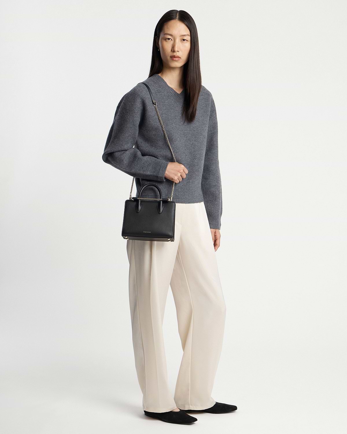 Model wearing grey jumper and cream trousers holding a black mini tote bag.