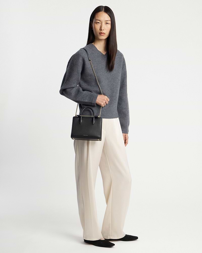 Model wearing grey jumper and cream trousers holding a black mini tote bag.