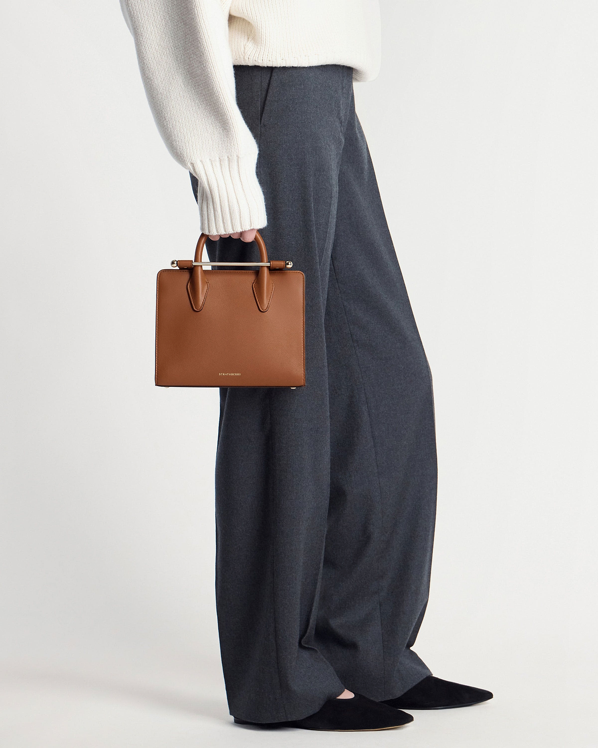 Strathberry chestnut mini tote held by a model wearing cream sweater and grey trousers