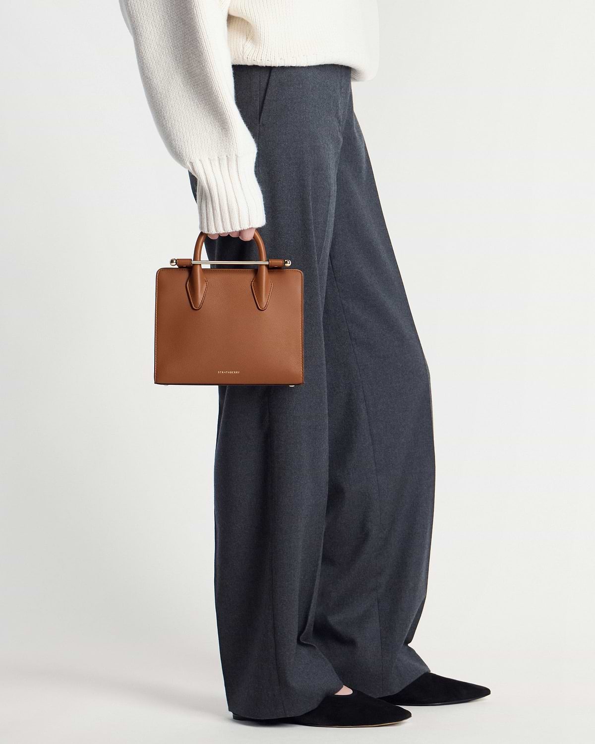 Strathberry chestnut mini tote held by a model wearing cream sweater and grey trousers