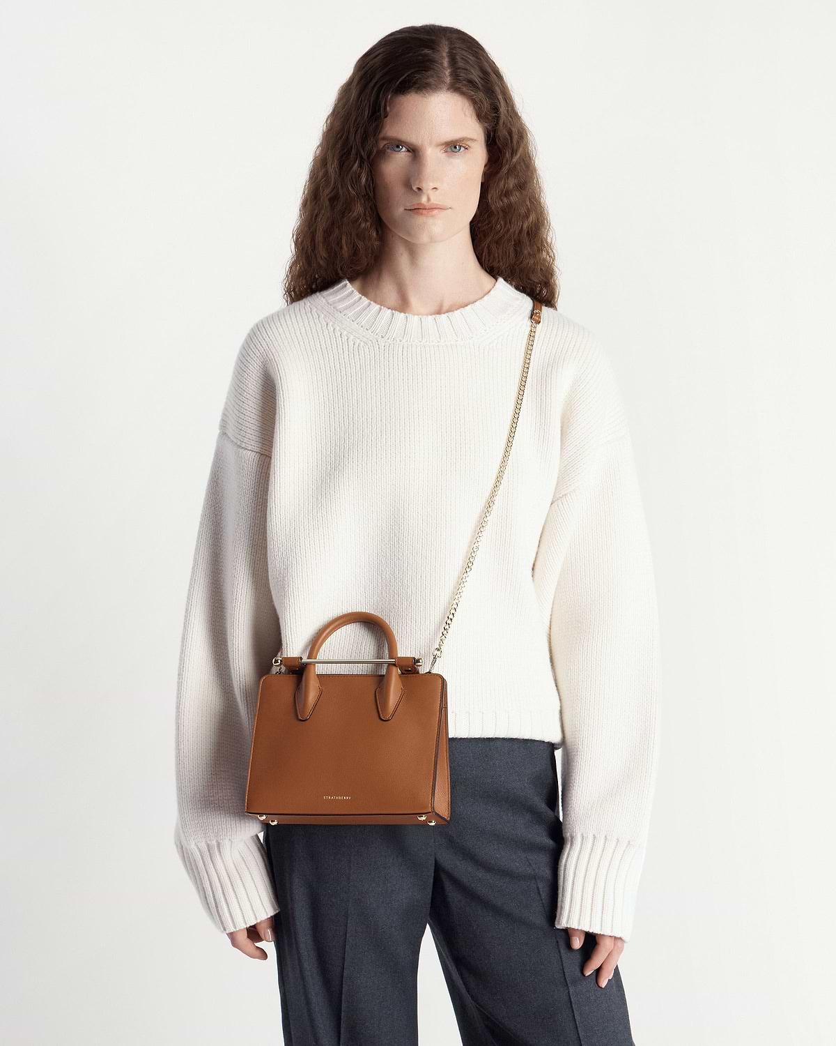 Model wearing chestnut Strathberry mini tote with chain strap over cream jumper.