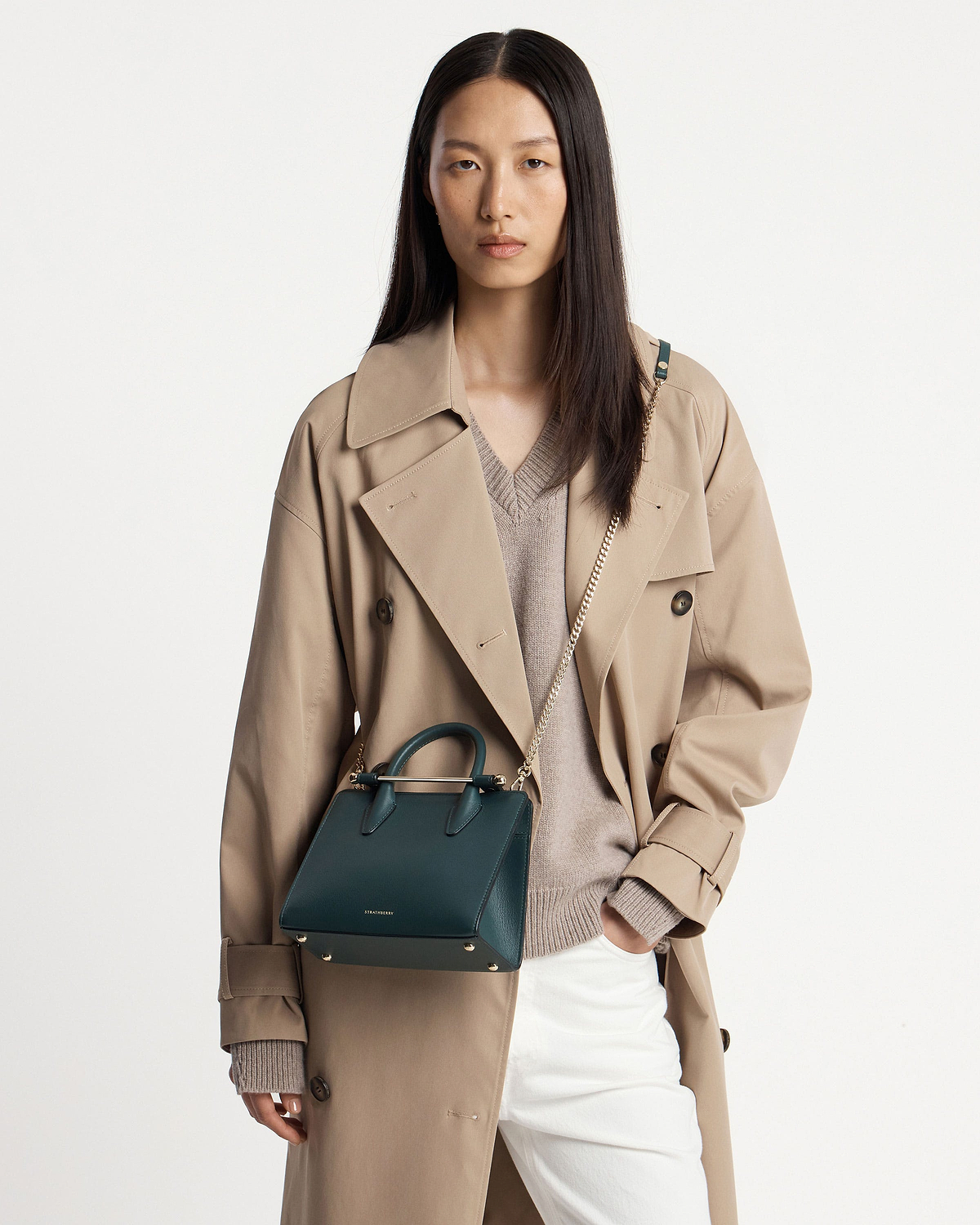 Model wearing a beige trench coat holding a bottle green Strathberry mini tote.