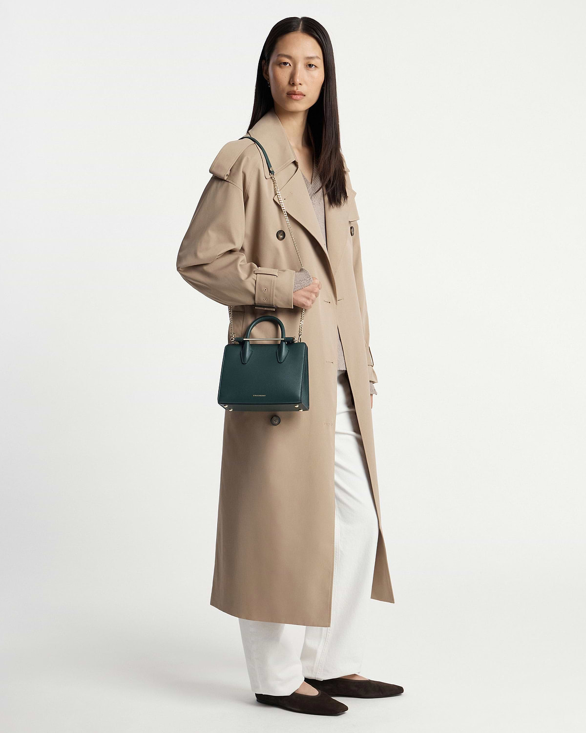 Model wearing beige trench coat holding bottle green Strathberry mini tote bag with chain strap.