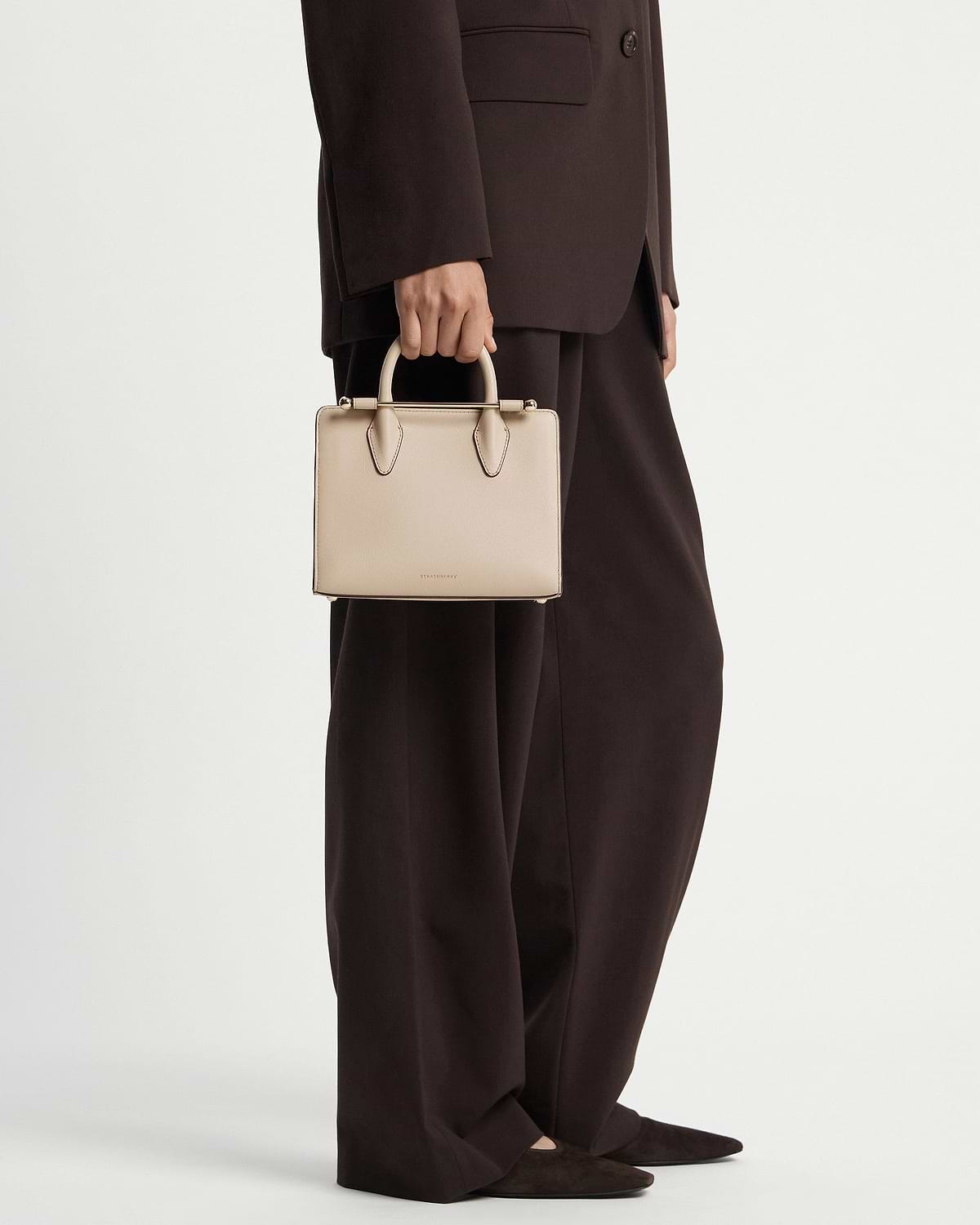 Model cropped below waist holding oat Strathberry mini tote with top handles.