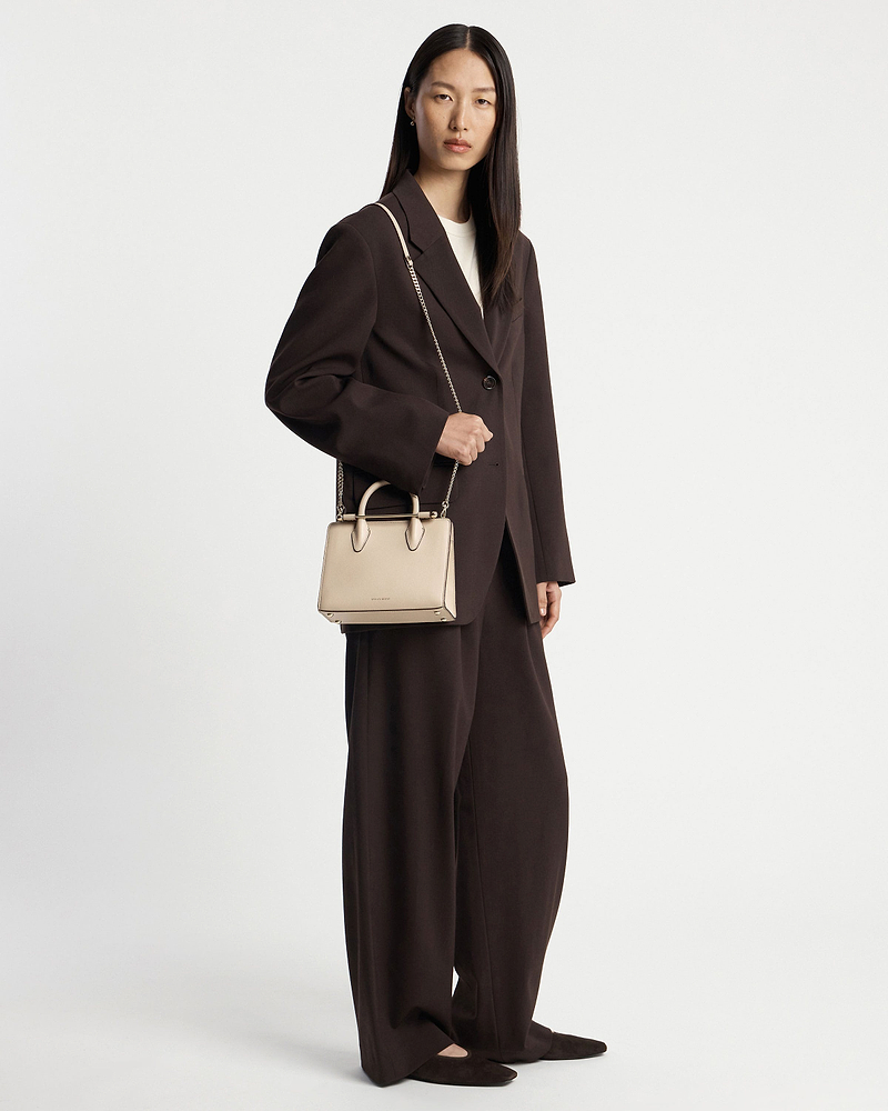 Model in dark brown suit holding oat mini tote by top handles and chain strap.