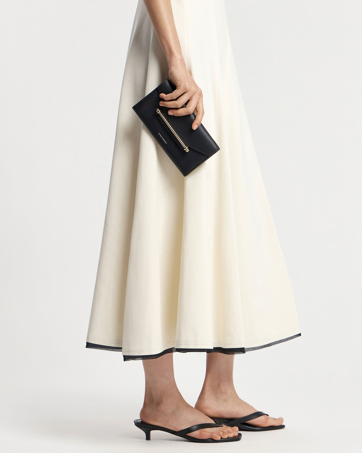 Model holding black leather envelope wallet with gold bar detail beside cream-coloured skirt.