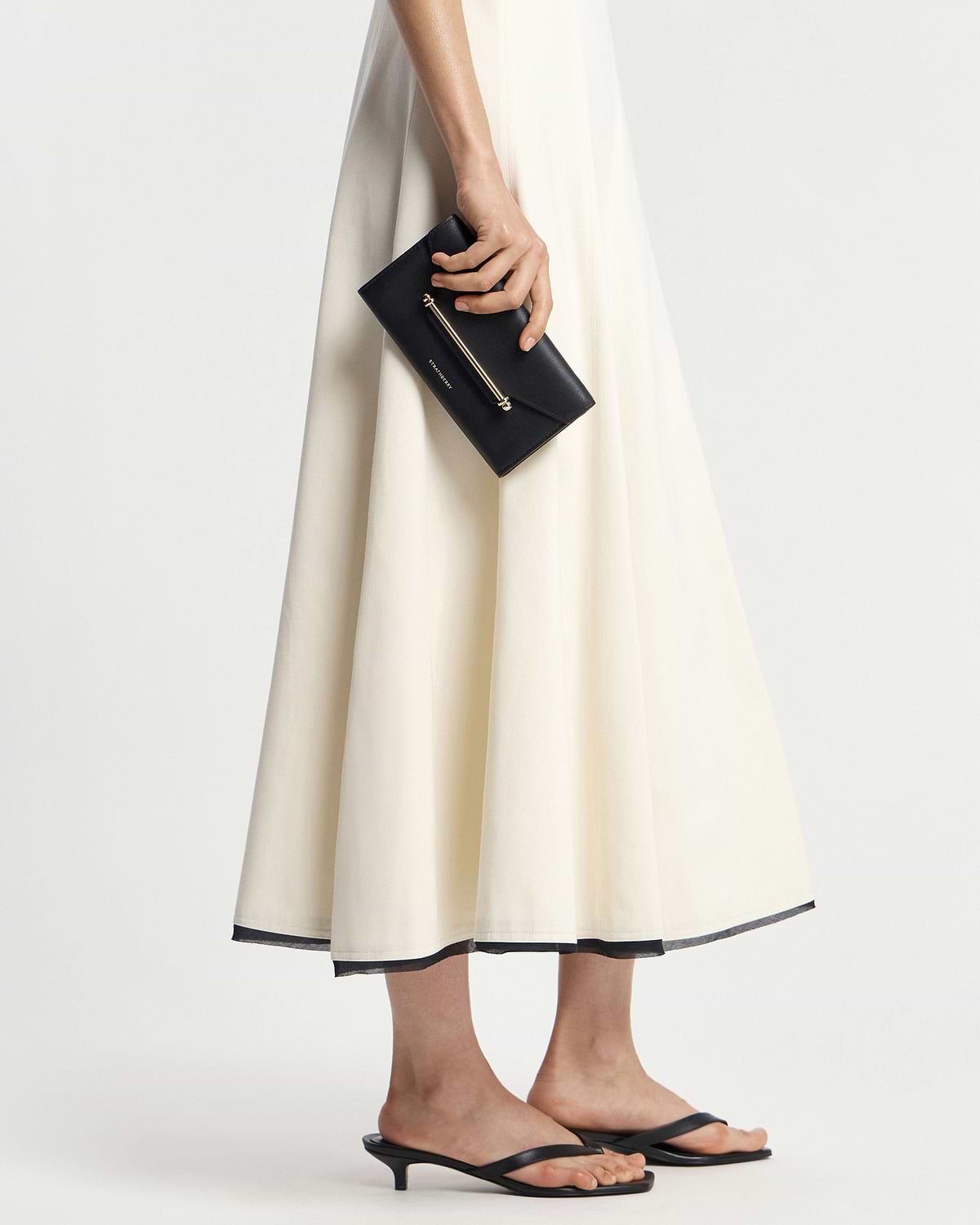 Model holding black leather envelope wallet with gold bar detail beside cream-coloured skirt.