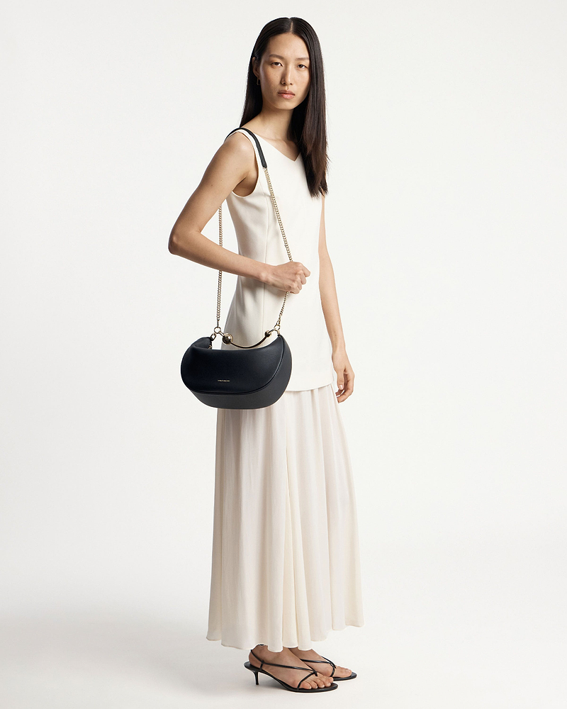 Model in white dress holding a black crescent moon bag with gold chain strap.