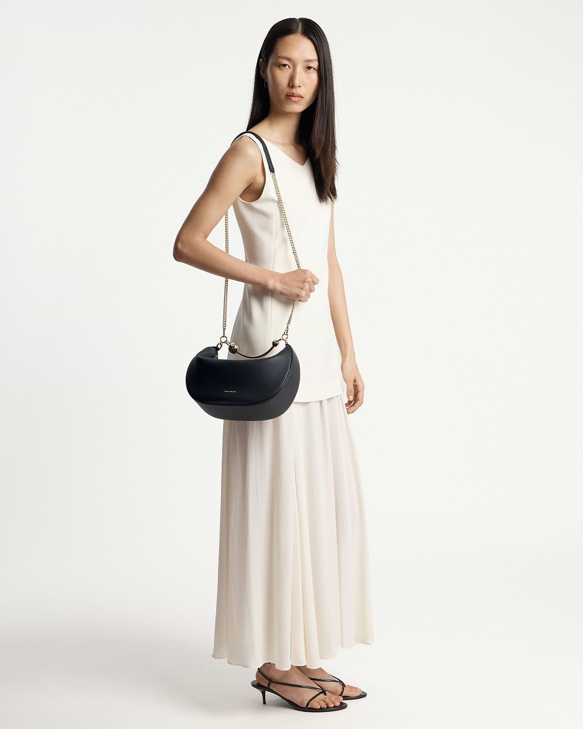 Model in white dress holding a black crescent moon bag with gold chain strap.