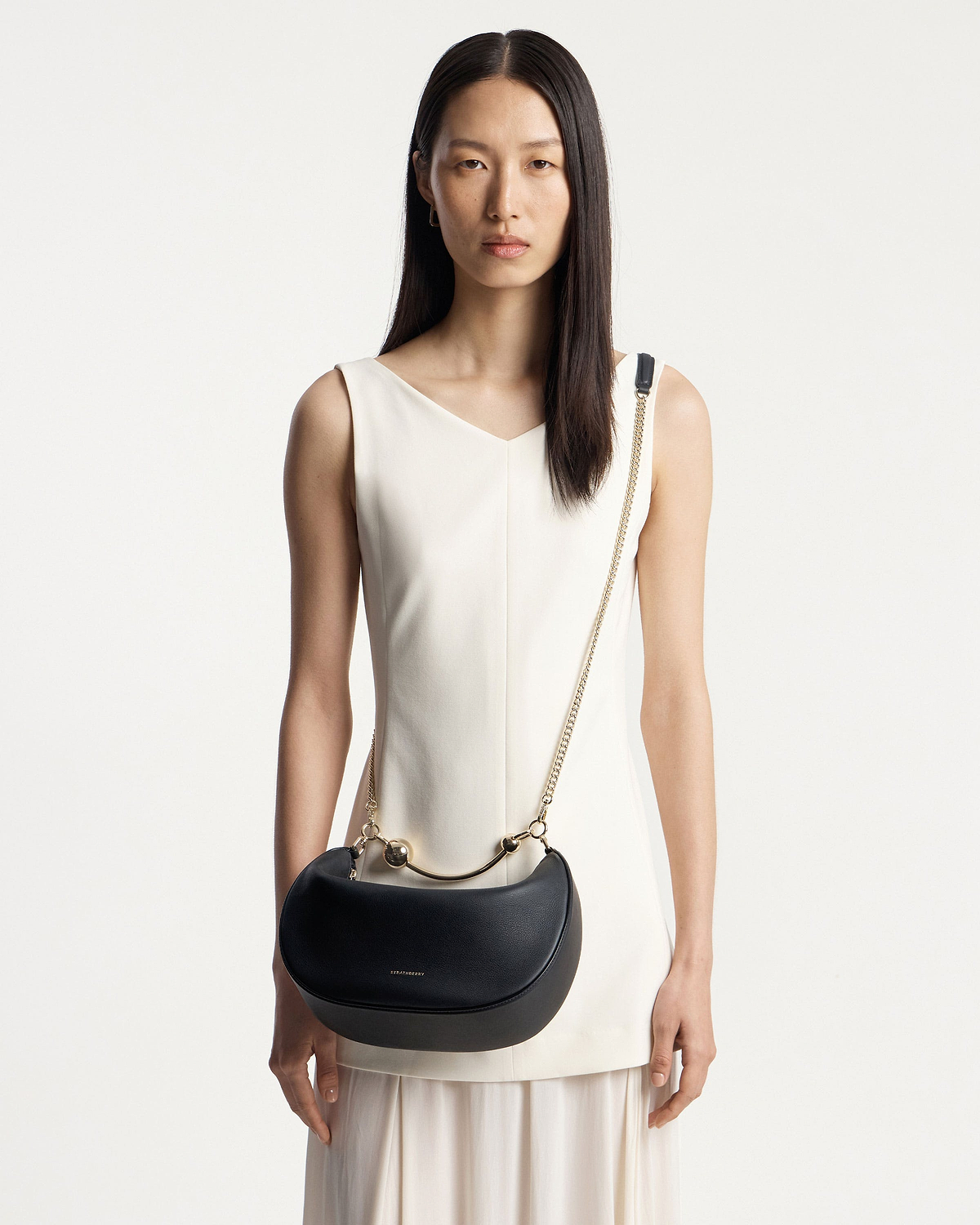 Model wearing white dress carrying black Strathberry Crescent Moon bag with gold chain strap.