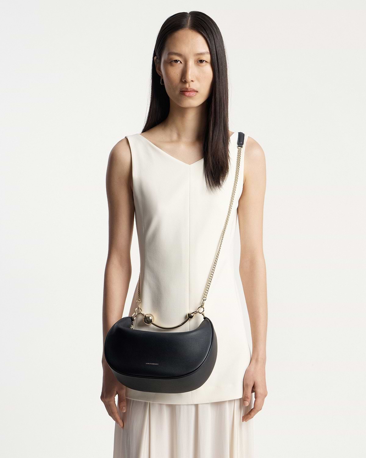 Model wearing white dress carrying black Strathberry Crescent Moon bag with gold chain strap.
