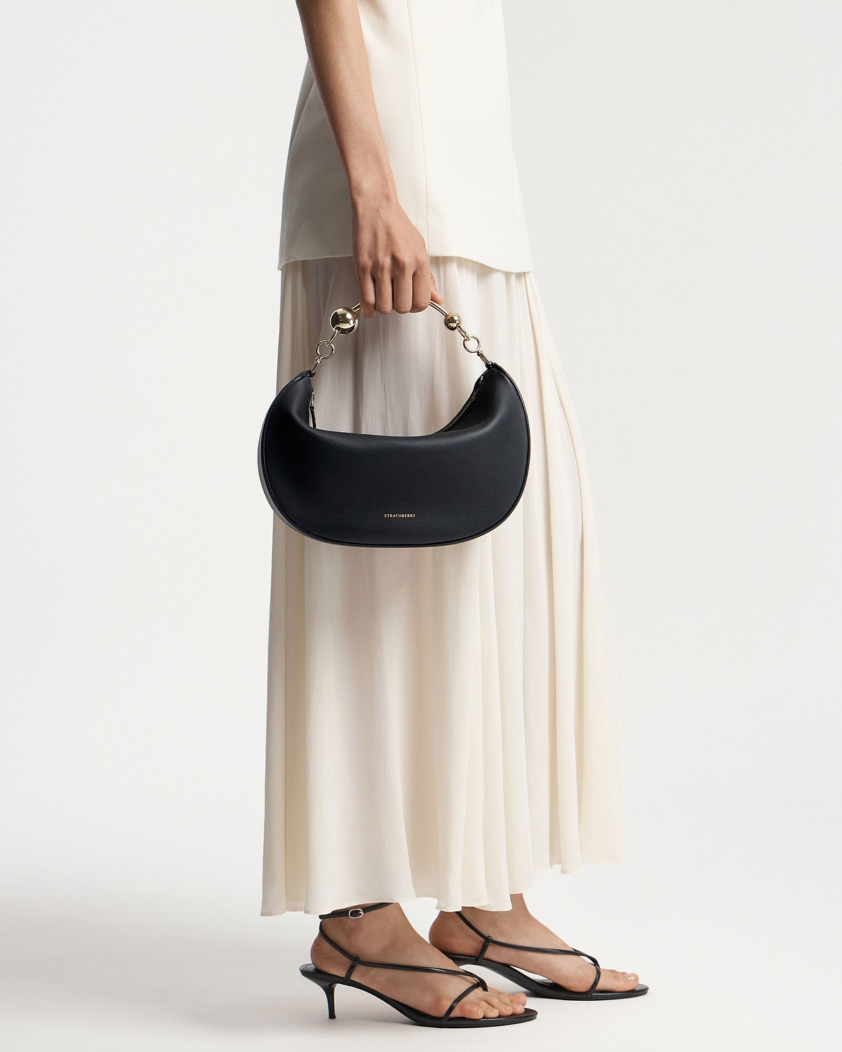 Model waist down holding a black Strathberry Crescent Moon bag with gold handle.