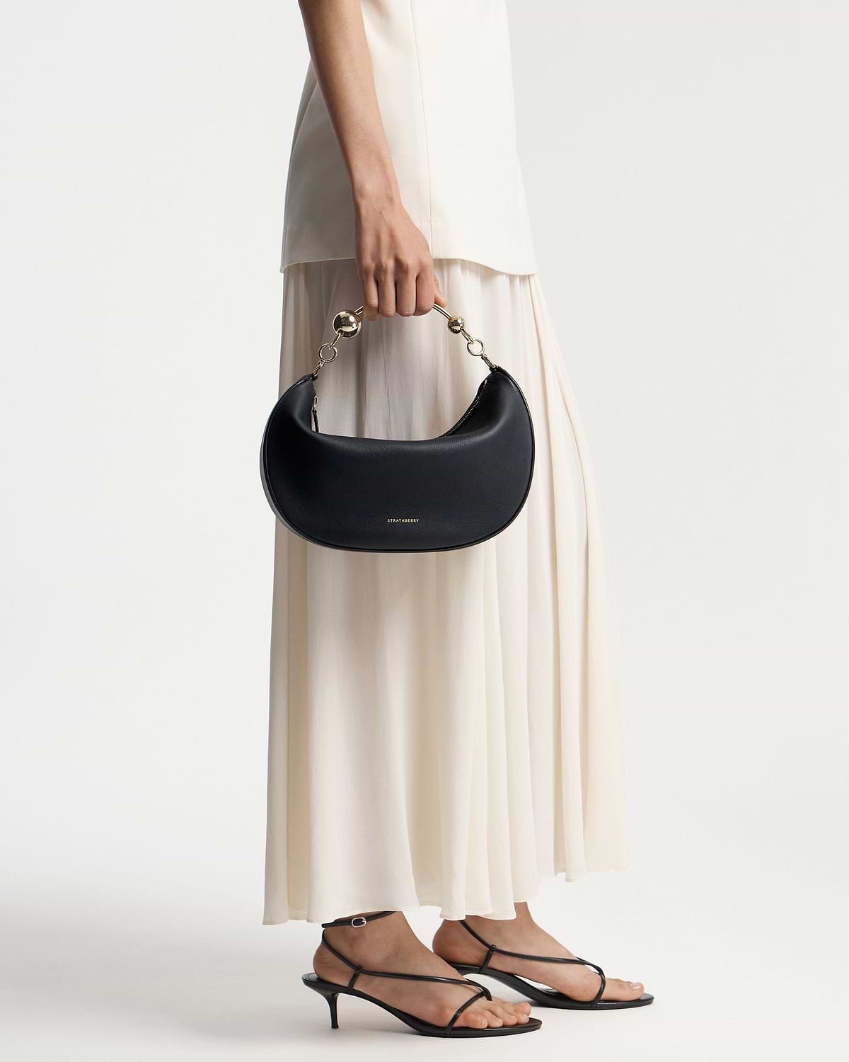 Model waist down holding a black Strathberry Crescent Moon bag with gold handle.