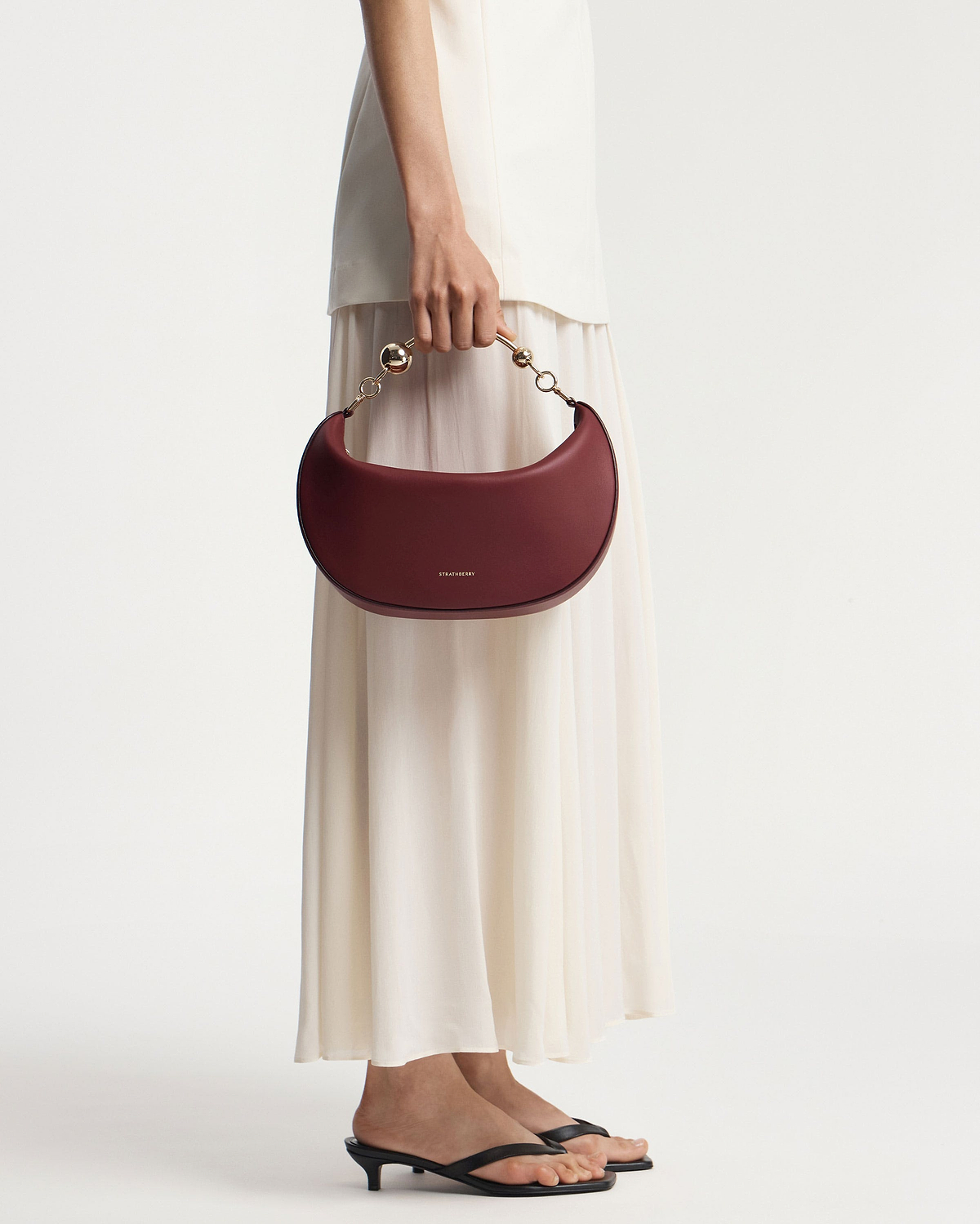 Model holding oxblood crescent moon bag with gold chain handle, Strathberry logo.
