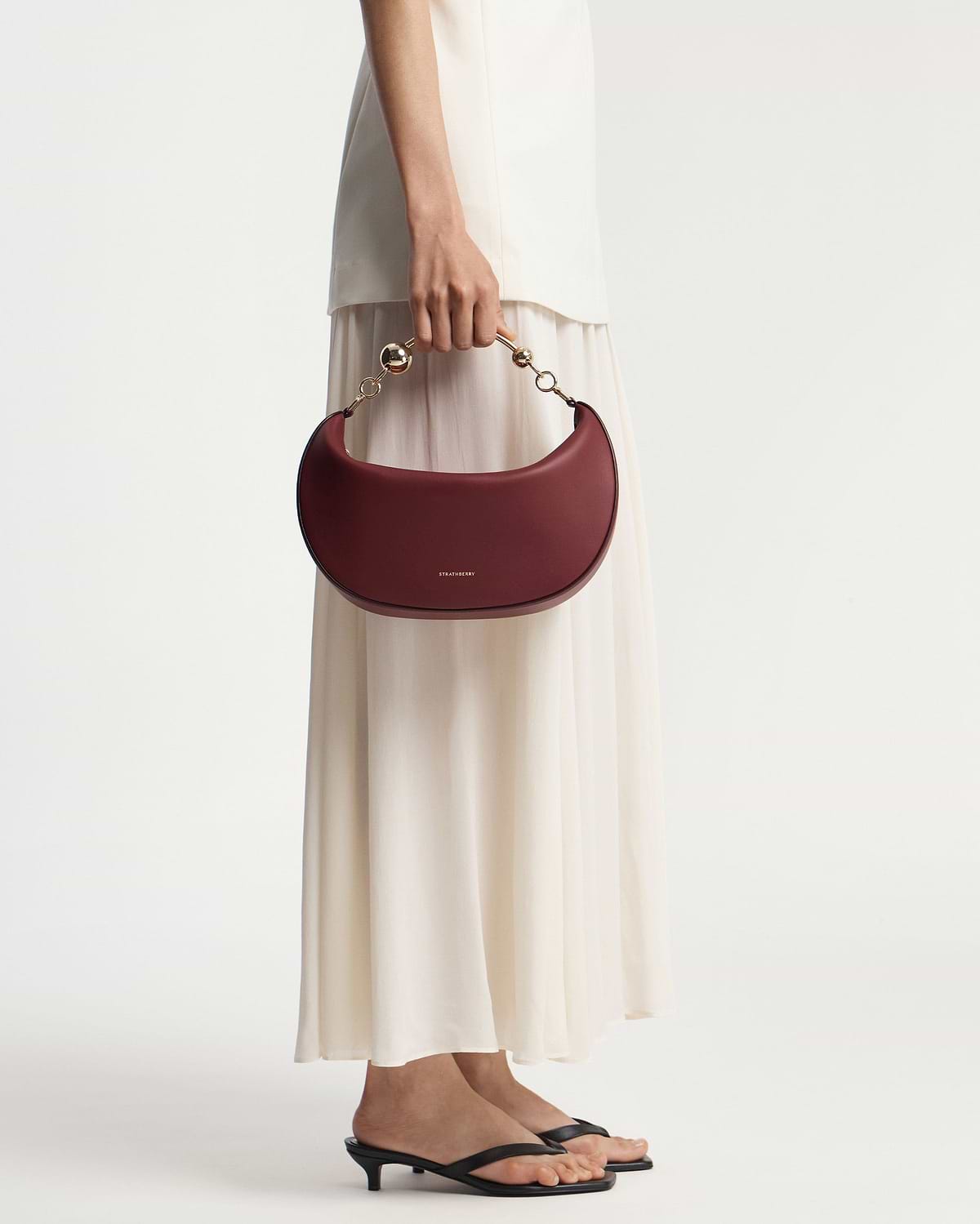 Model holding oxblood crescent moon bag with gold chain handle, Strathberry logo.