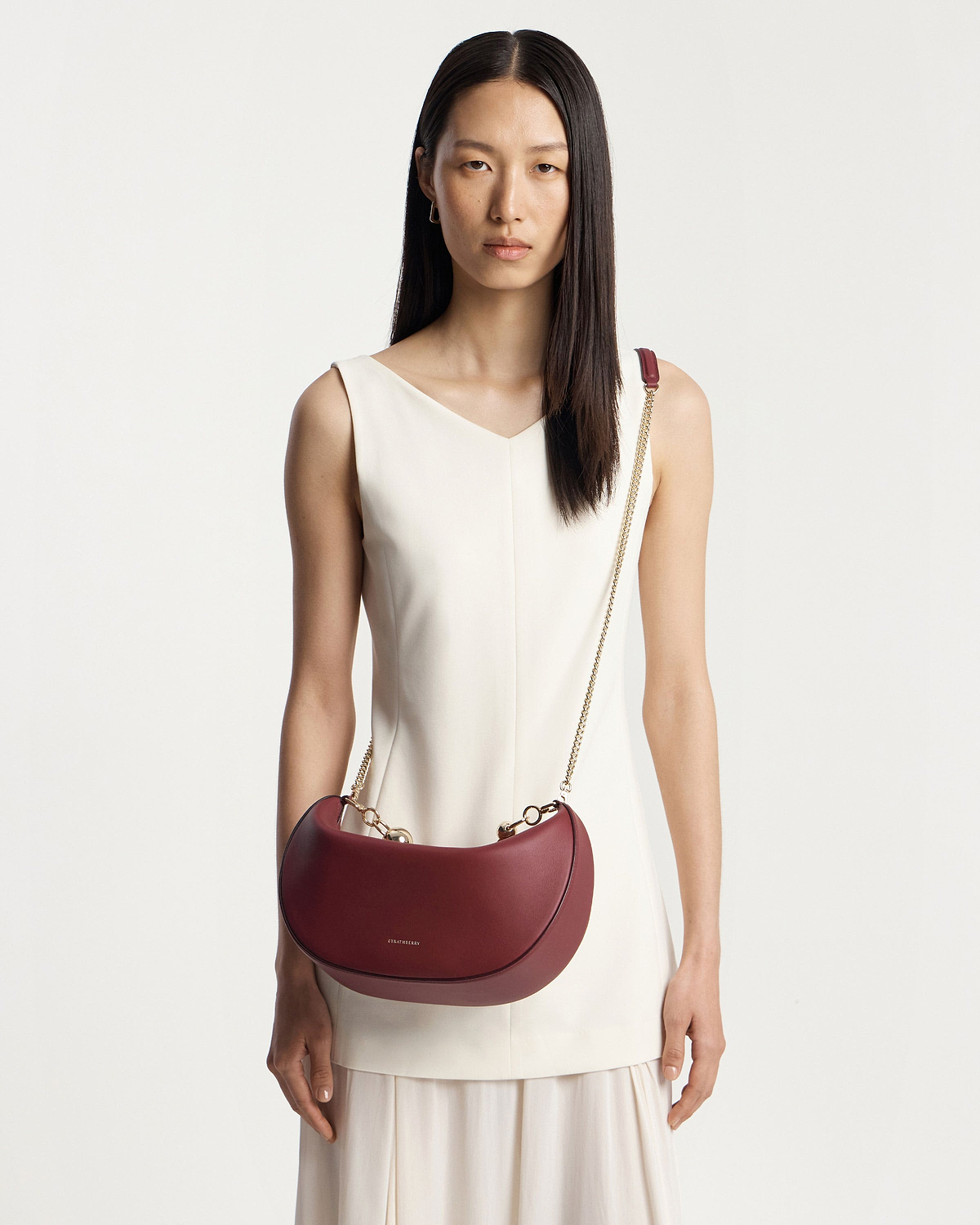 Model wearing white sleeveless dress carrying oxblood crescent moon bag with gold chain strap.