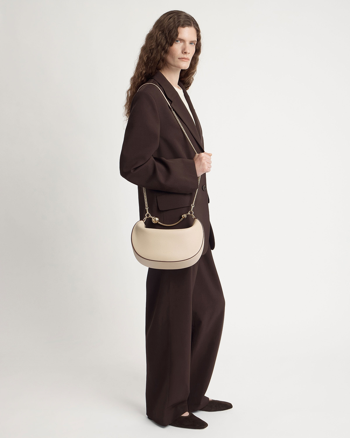 Model in dark brown suit carries oat crescent moon shoulder bag with gold chain.