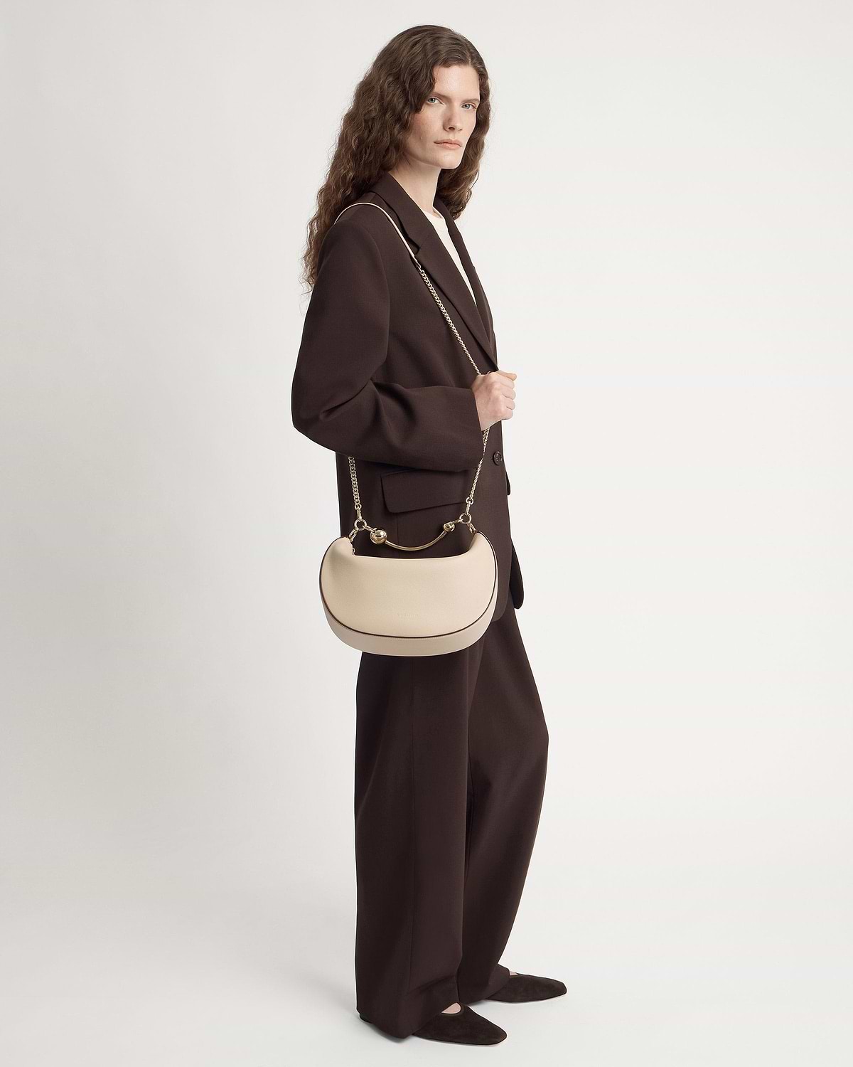 Model in dark brown suit carries oat crescent moon shoulder bag with gold chain.