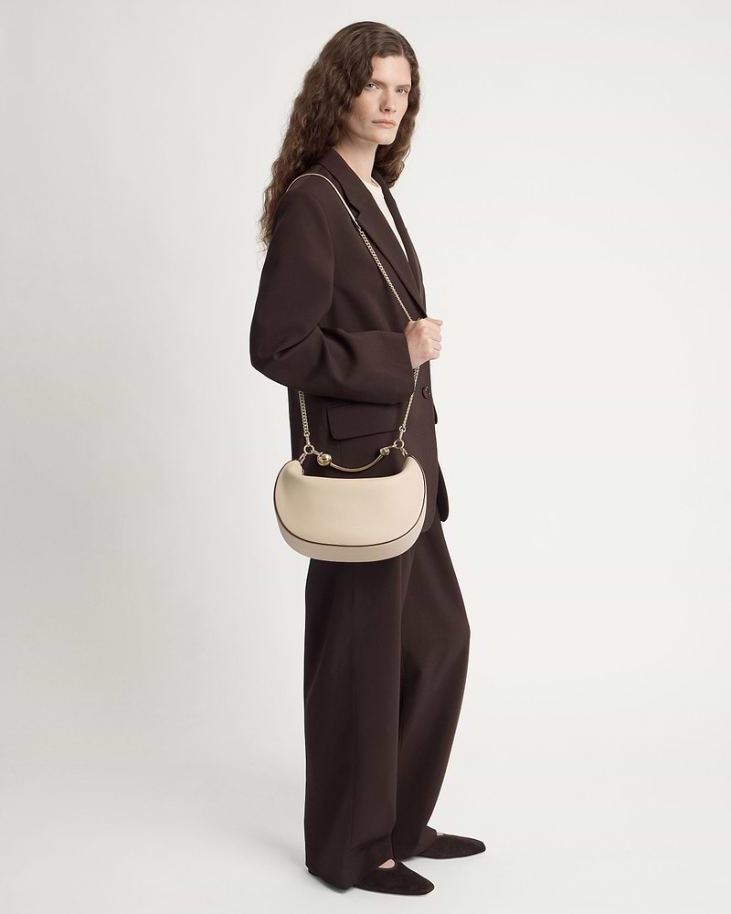 Model in dark brown suit carries oat crescent moon shoulder bag with gold chain.