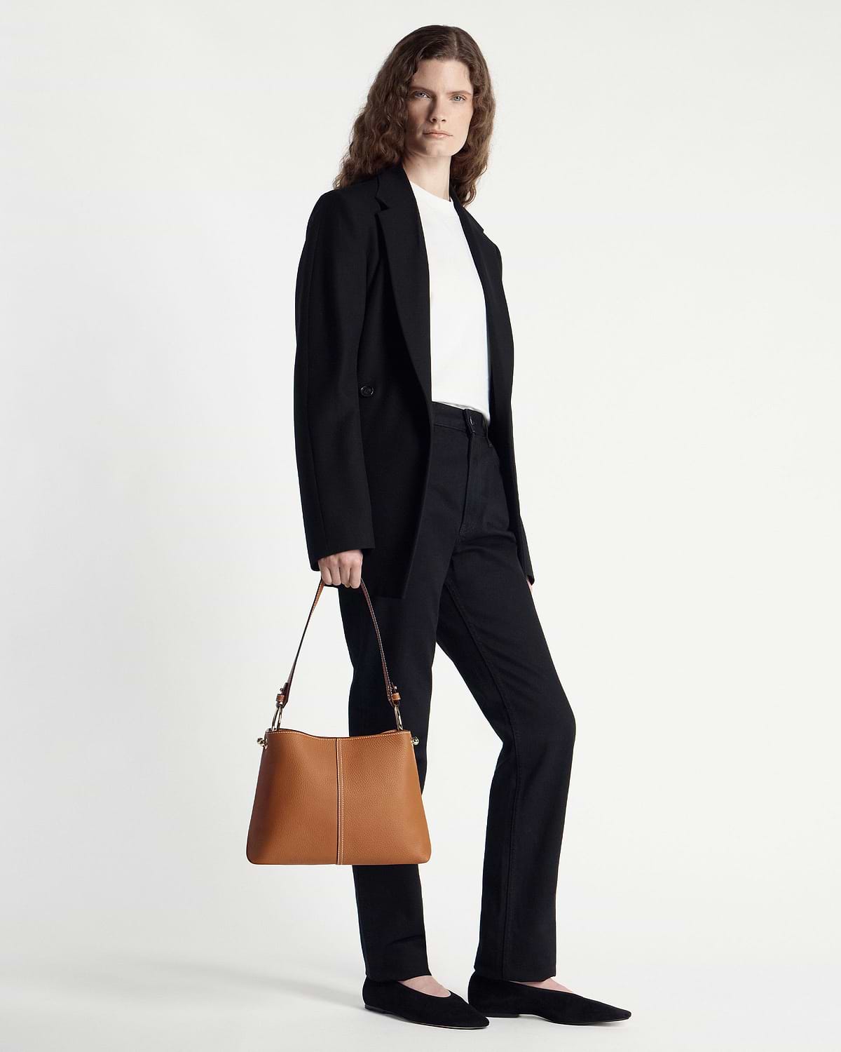 Model in a black blazer and trousers holding a tan hobo bag at her side.