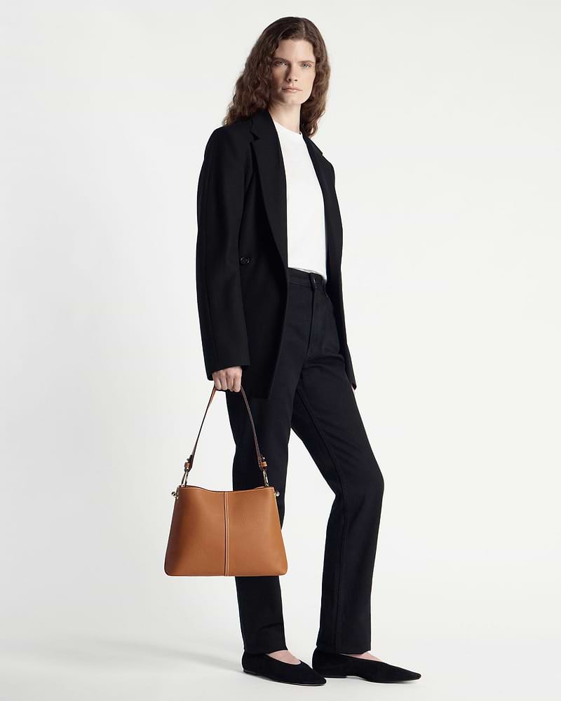 Model in a black blazer and trousers holding a tan hobo bag at her side.