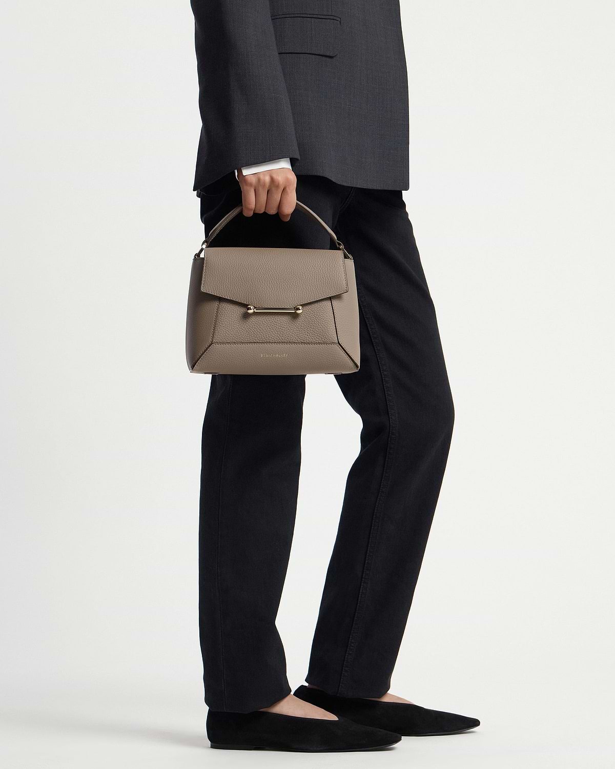 Model holding taupe mosaic bag by top handle, grey blazer and black trousers visible.