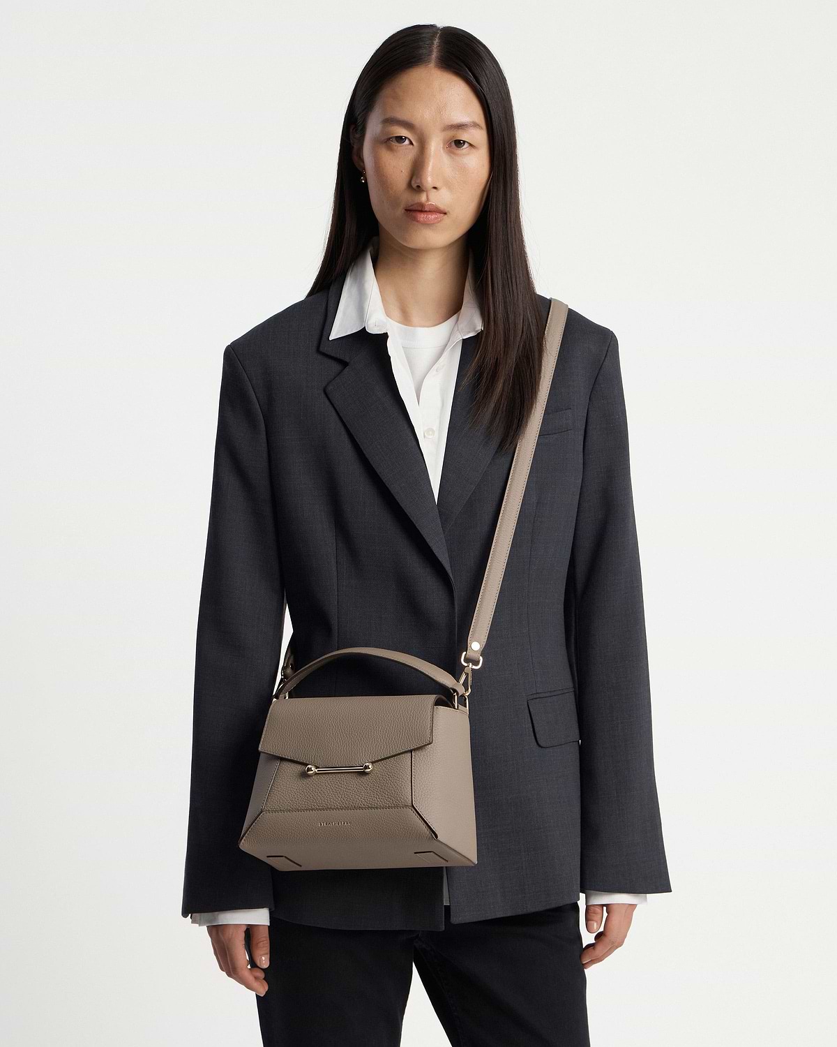 Model wearing dark blazer carrying taupe mosaic bag with top handle and shoulder strap.