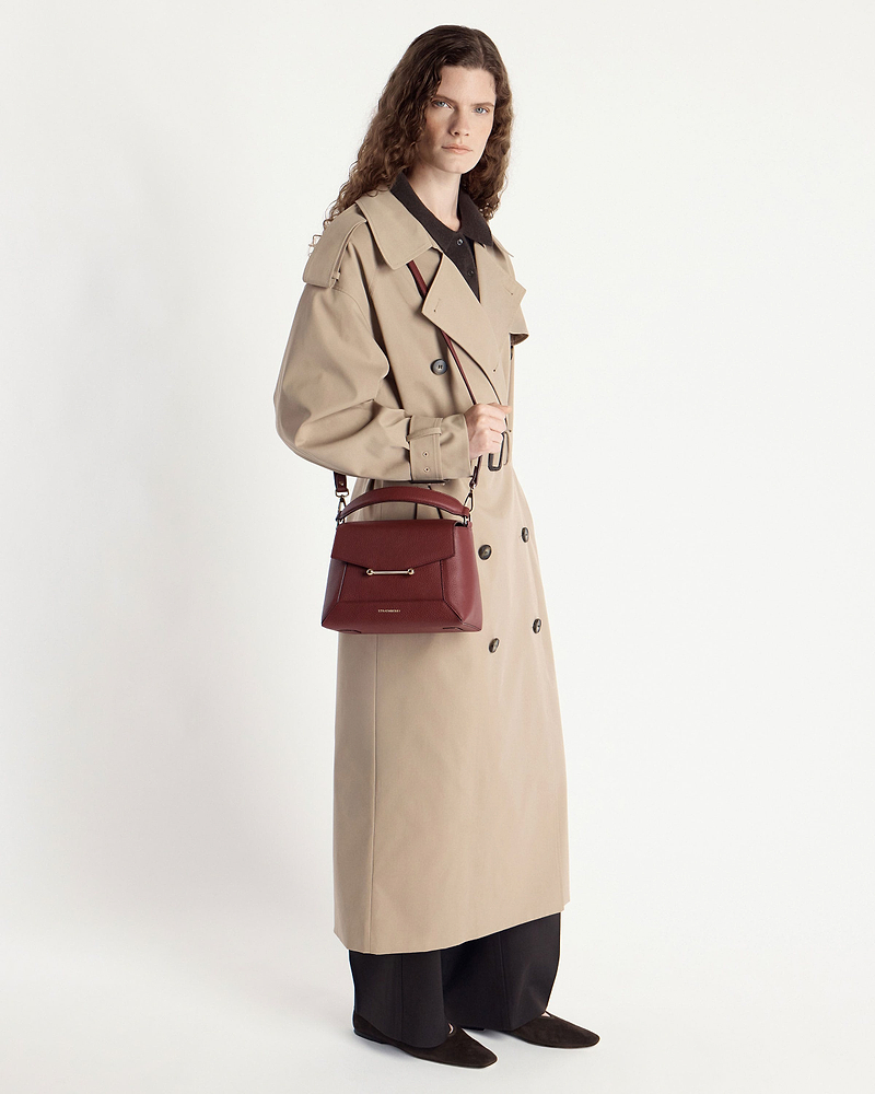Model in beige trench coat holding an oxblood mosaic bag by the handle.