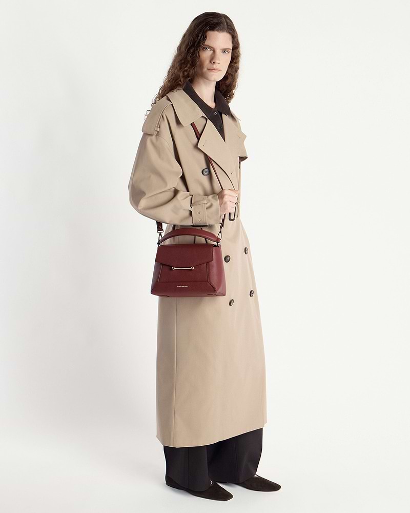 Model in beige trench coat holding an oxblood mosaic bag by the handle.
