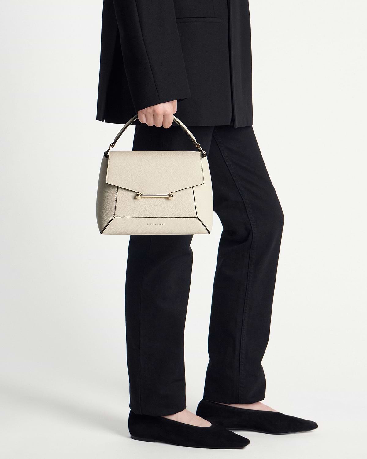 Model holding a vanilla Strathberry Mosaic bag by its handle, wearing black trousers and flats.