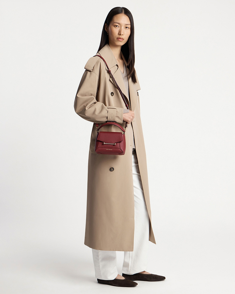 Model wearing a beige trench coat carrying a small oxblood crossbody bag at hip.