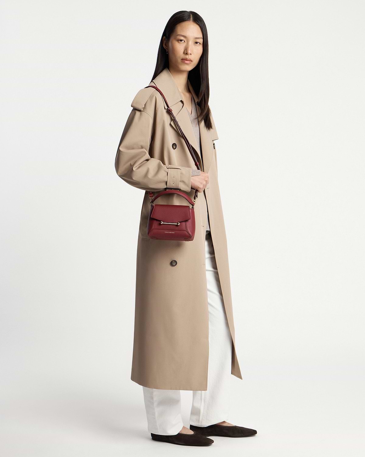 Model wearing a beige trench coat carrying a small oxblood crossbody bag at hip.