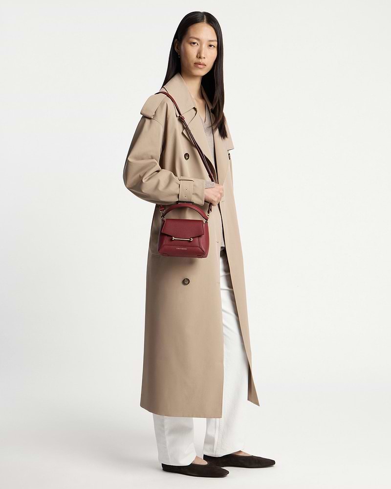 Model wearing a beige trench coat carrying a small oxblood crossbody bag at hip.