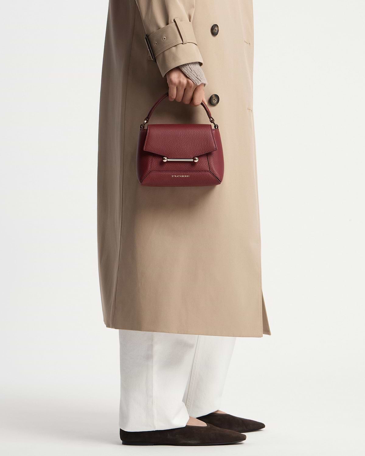 Oxblood Mosaic Nano top-handle bag by Strathberry held against beige trench coat.