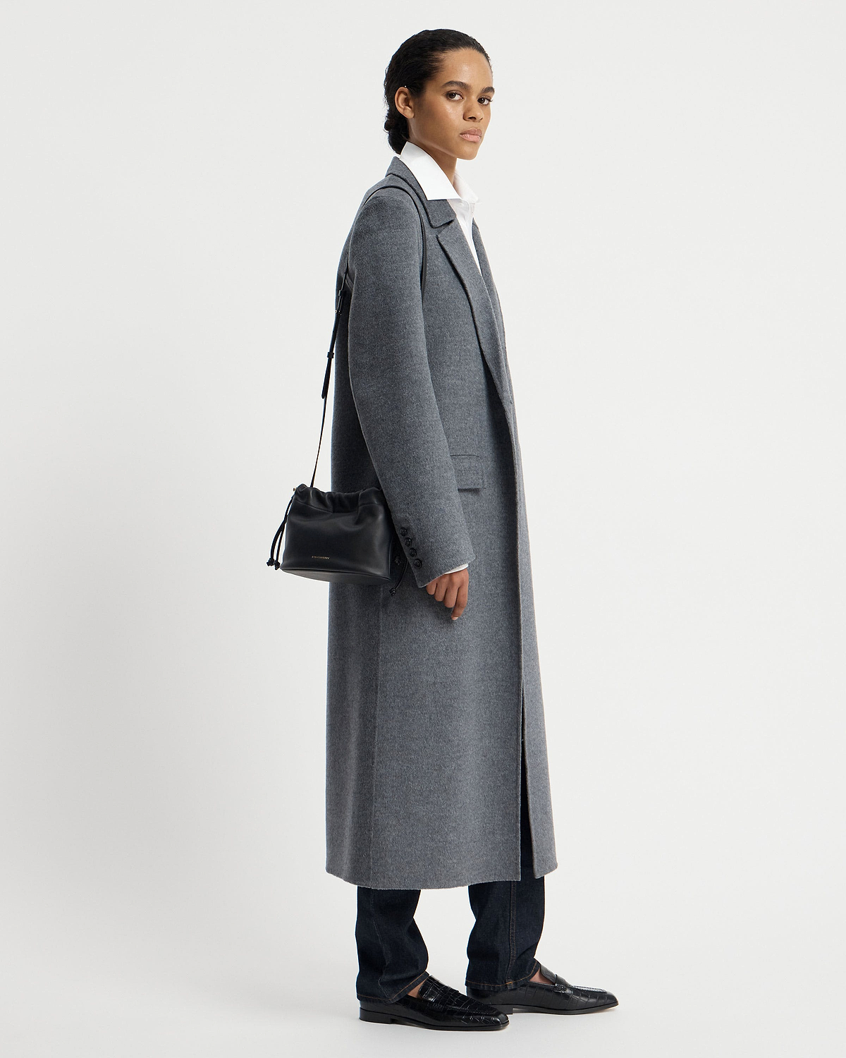 Model in grey coat carrying a black drawstring shoulder bag at her side.