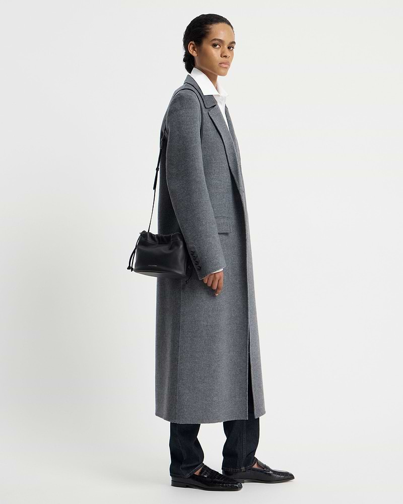 Model in grey coat carrying a black drawstring shoulder bag at her side.