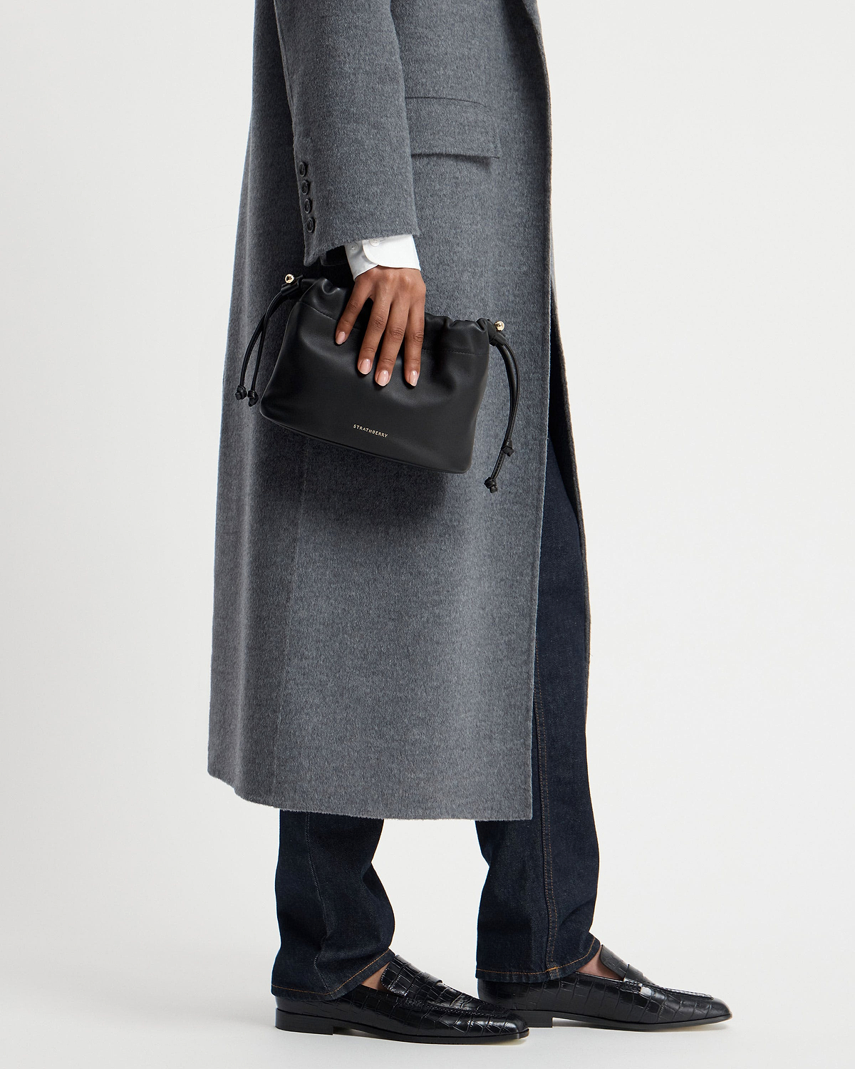Hand holding black Strathberry Charlotte Drawstring bag against grey coat.
