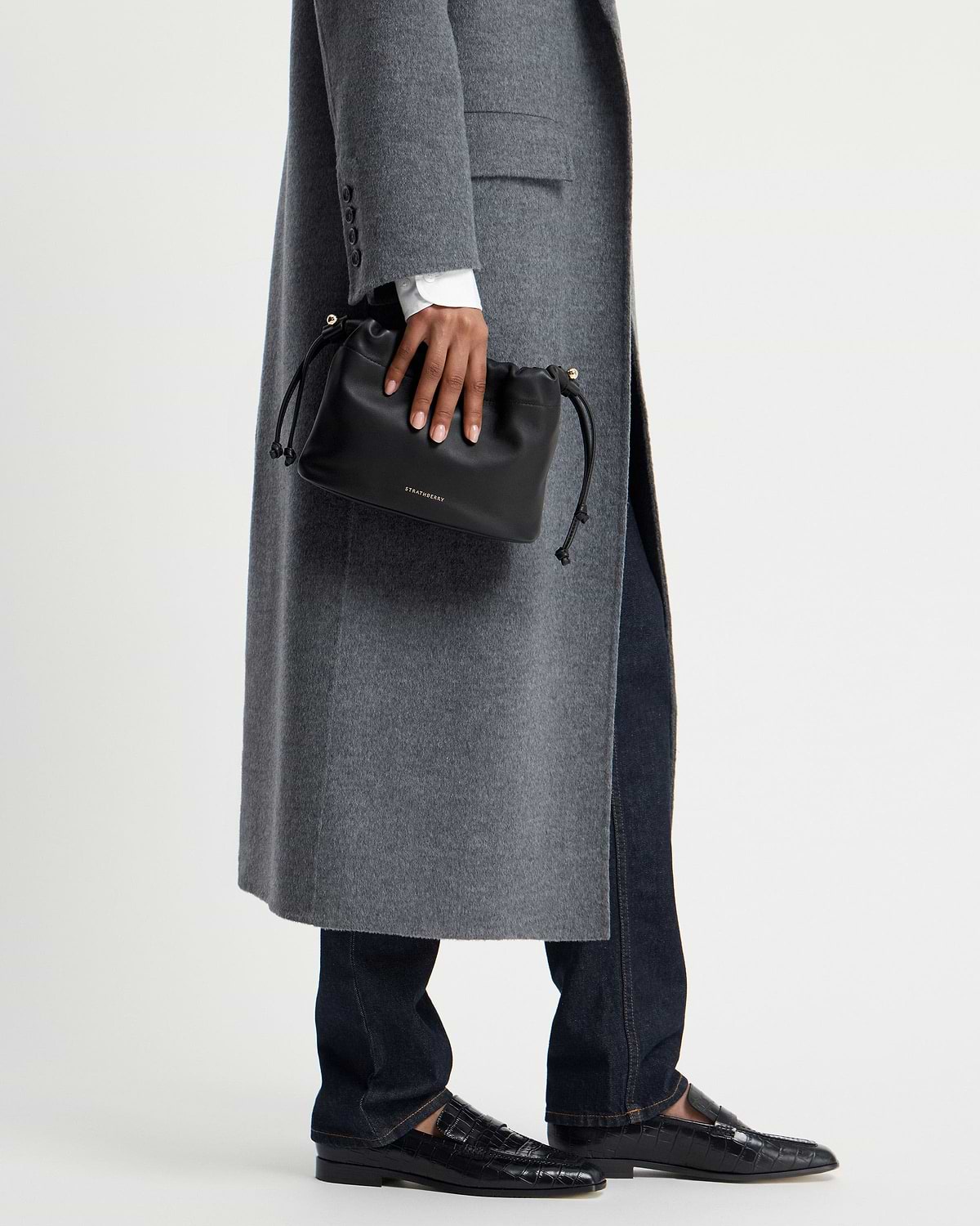 Hand holding black Strathberry Charlotte Drawstring bag against grey coat.